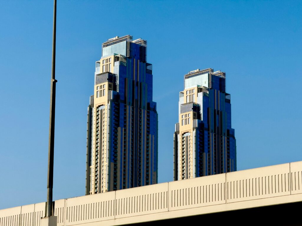 Modern twin skyscrapers rise against a cloudless blue sky, viewed over a bridge.