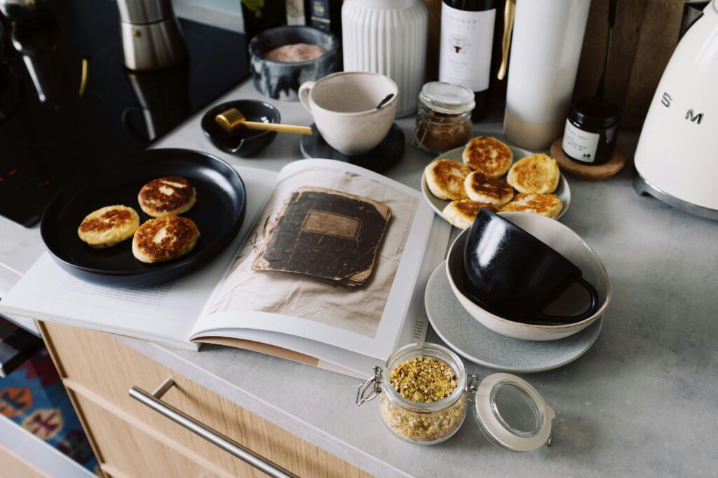 High angle of cookbook and cooked fritters on plates placed on table near utensils and ingredients in cozy kitchen at home