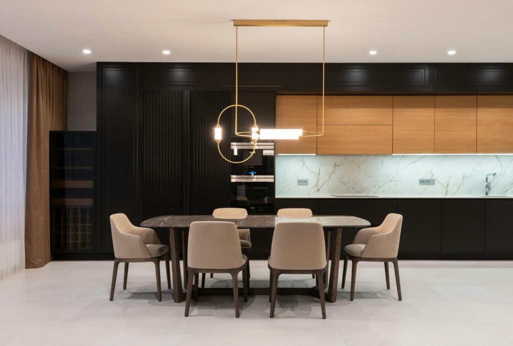 Interior of modern bright house with dining table with chairs under pendant lamp against open plan kitchen with black and wood cabinets