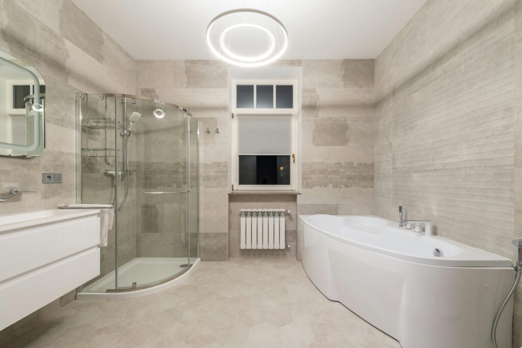 White bathtub near sink with mirror and glass shower cabin placed in stylish modern bathroom with window and glowing chandelier